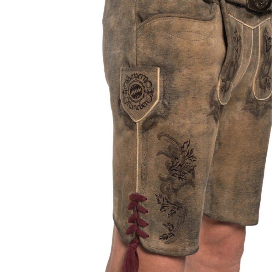 Close-up of Bavarian Heritage lederhosen left leg showing Bayern München crest patch and detailed floral embroidery with red lacing tassel