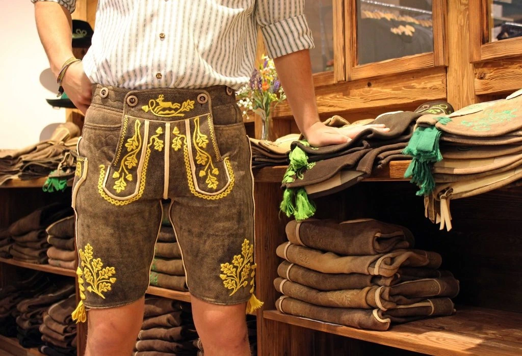Man wearing traditional Bavarian lederhosen with intricate yellow embroidery standing in front of a shelf stacked with similar garments.
