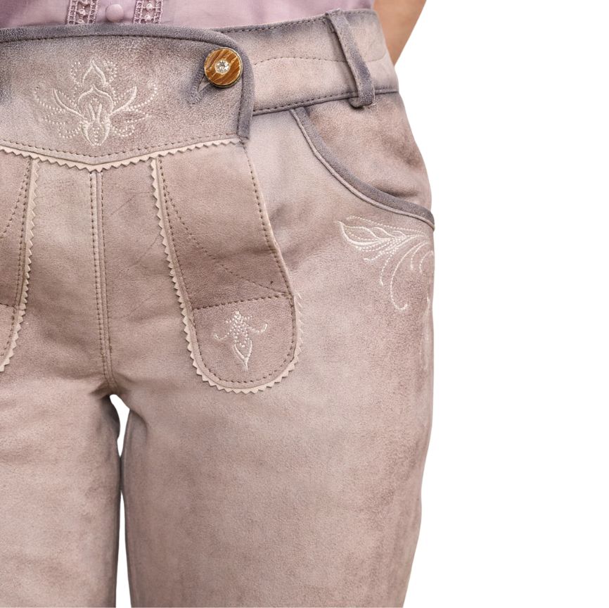 Close-up of beige leather lederhosen showing white embroidered motifs on the front flap and pocket area with a decorative wooden button.